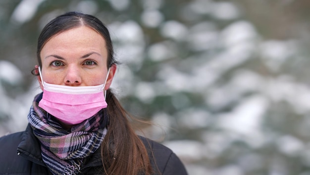 Junge Frau in Winterjacke mit rosafarbener Einweg-Gesichtsvirusmaske zum einmaligen Gebrauch unsachgemäß - Nase nicht bedeckt, Platz für Text (verschwommener Hintergrund schneebedeckte Bäume) rechte Seite