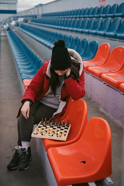 Junge Frau in Winterkleidung spielt Schach und sitzt allein auf Stadiontribünen Frau in schwarzer Mütze mit Schach im Sportstadion bei bewölktem Wetter