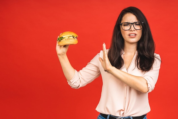 Junge Frau isoliert auf rotem Hintergrund Studioporträt Richtige Ernährung oder amerikanisches klassisches Fast-Food-Konzept Mock-up-Kopienbereich Zeigt Stop-Geste mit Palme zum Burger