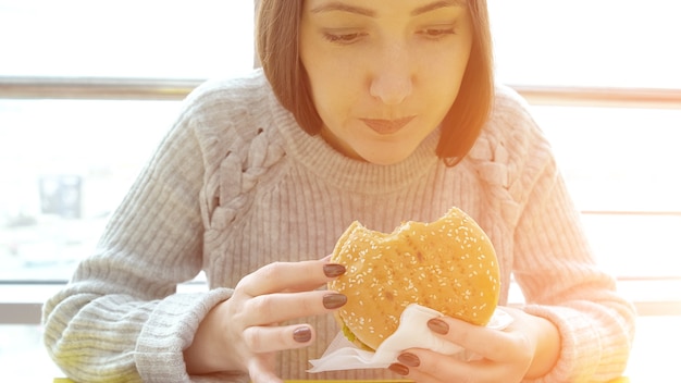 Junge Frau isst einen Burger, Sonnenlicht. Schädliche fetthaltige Lebensmittel.