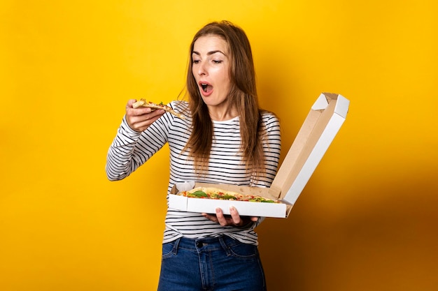 Junge Frau isst frische heiße Pizza mit Verpackung auf gelbem Hintergrund