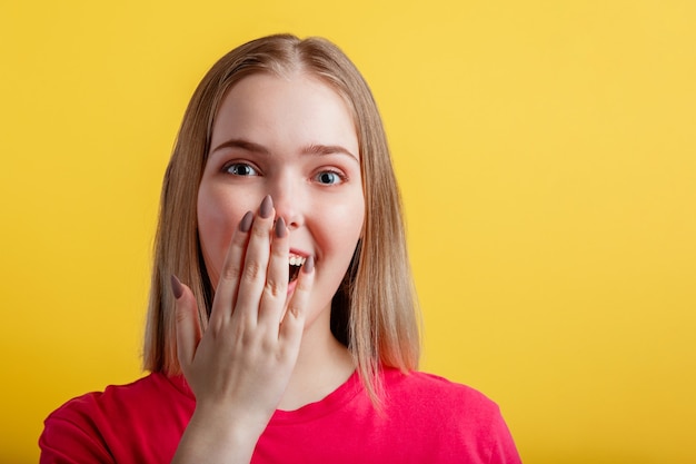 Junge Frau ist angenehm überrascht. Blondes Mädchen im Teenageralter bedeckte ihren offenen Mund mit der Hand überrascht. Positives emotionales Porträt der jungen Frau auf der gelben Wand der Farbe mit Kopienraum.