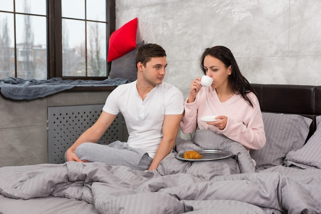 Junge Frau ist gerade aufgewacht, hat eine Tasse Kaffee getrunken und ihr Frühstück im Bett mit ihrem Freund in der Nähe des Sitzens im Schlafanzug gegessen