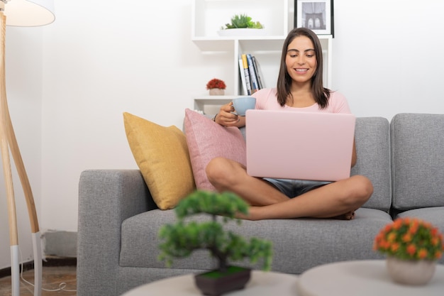 Junge Frau lächelt glücklich und macht einen Videoanruf mit ihren Freunden auf ihrem Laptop, während sie Kaffee trinkt