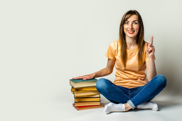 Junge Frau lächelt und macht eine Handbewegung, die ihre Hand auf einem Stapel Bücher hält, die auf dem Boden auf einem hellen Raum sitzen