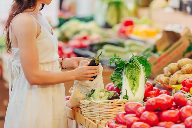 Junge Frau legt Obst und Gemüse in Baumwollbeutel auf dem Lebensmittelmarkt. Wiederverwendbare Öko-Tasche zum Einkaufen. Zero-Waste-Konzept.