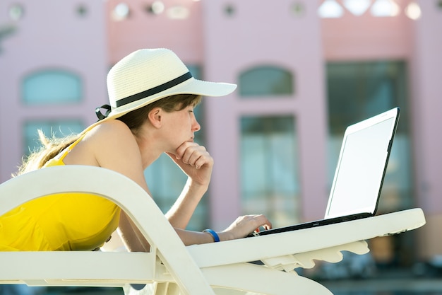 Junge Frau liegt auf Strandkorb, der an Computer-Laptop arbeitet, der mit drahtlosem Internet verbunden ist, das Text auf Schlüsseln im Sommerresort tippt.