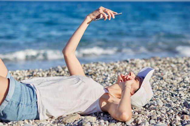 Junge Frau liegt in einem Hut mit einem Telefon am Strand in der Nähe des Meeres und macht ein Selfie Ruhendes Reise- und Entspannungskonzept Kopierbereich Hochwertiges Foto