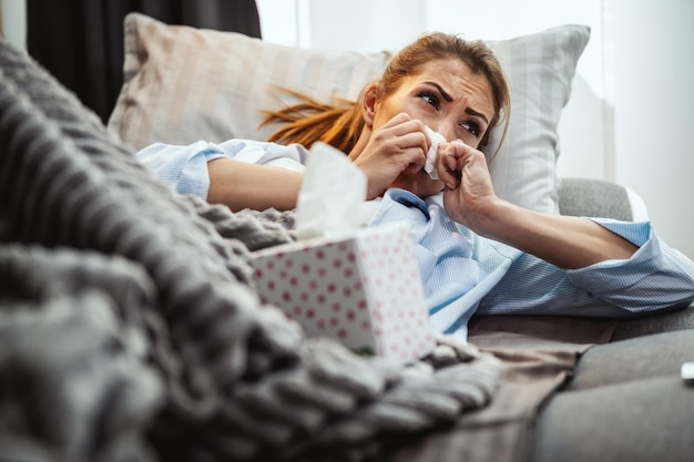 Junge Frau liegt krank auf der heimischen Couch und schnäuzt sich mit Papiertaschentuch die Nase.