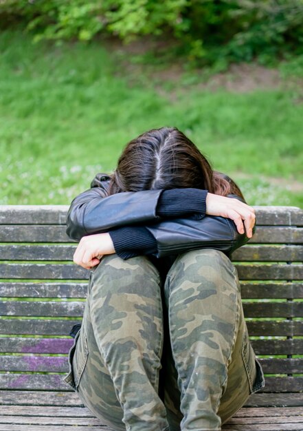 Junge Frau liegt traurig und deprimiert auf einer Parkbank, die Arme und den Kopf auf den Knien