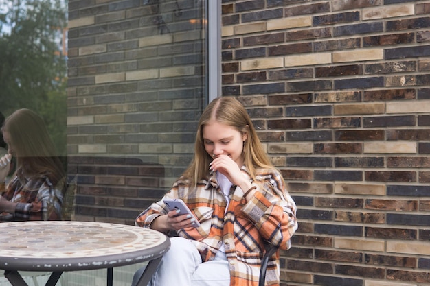 Junge Frau liest am Telefon eine lustige Nachricht, während sie an einem Tisch in der Cafeteria im Freien sitzt