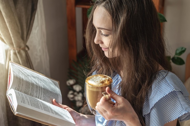 Junge Frau liest ein Buch und trinkt Kaffee am Fenster sitzend Konzept der morgendlichen Freude, Freizeit, angenehmer Zeitvertreib und Entspannung