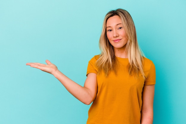 Junge Frau lokalisiert auf blauer Wand, die einen Kopienraum auf einer Handfläche zeigt und eine andere Hand auf Taille hält