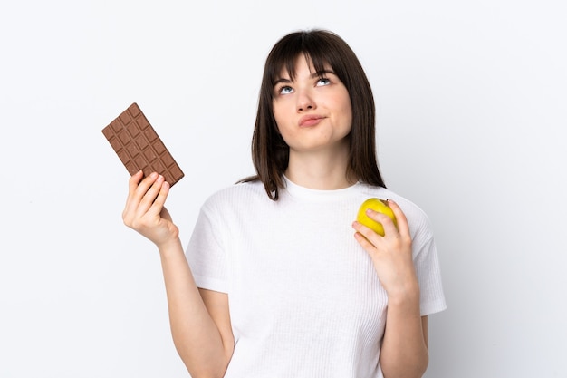 Junge Frau lokalisiert auf Weiß, das Zweifel hat, während sie eine Schokoladentafel in einer Hand und einen Apfel in der anderen nimmt
