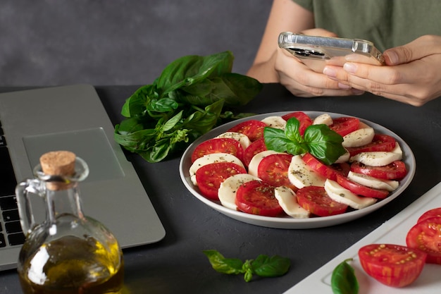 Junge Frau macht einen Caprese-Salat nach einem Rezept aus dem Internet und fotografiert das Ergebnis der Arbeit mit dem Smartphone.