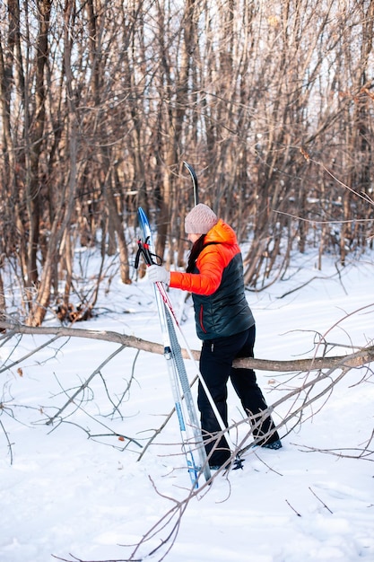 Junge Frau macht Sport im Wald Brünette im Winter-Trainingsanzug geht die verschneite Straße entlang und hält Skistöcke und Skier in der Seitenansicht der Hände