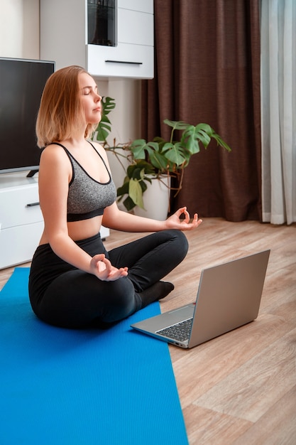 Junge Frau macht Yoga-Übungen im Wohnzimmer mit Laptop-Teenager-Mädchen in Sportkleidung machen Remote-Yoga-Kurse im Lotussitz während der Meditation zu Hause, um Stress-Wellness zu reduzieren