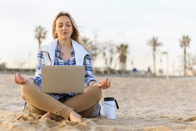 Foto junge frau meditiert an einem leeren strand mit laptop und kaffeetasse