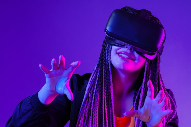 Junge Frau mit Afro-Zöpfen mit VR-Brille auf dunkelviolettem Neonhintergrund