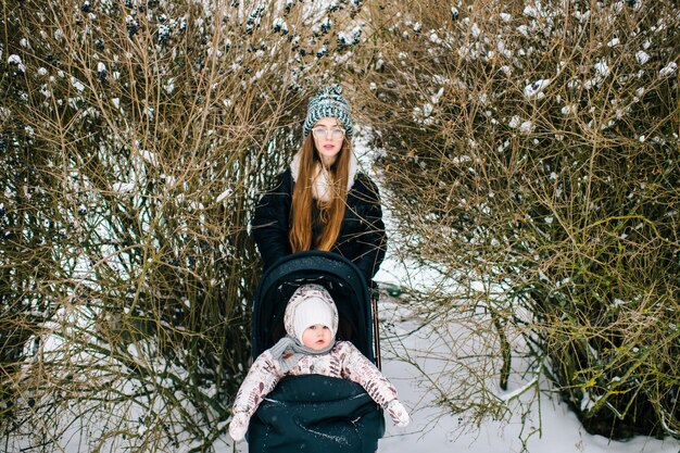 Junge Frau mit Baby im Kinderwagen in den Büschen am Wintertag.