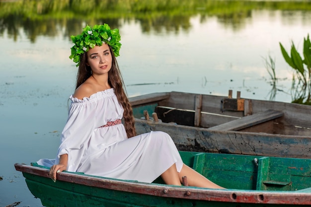 Junge Frau mit Blumenkranz auf dem Kopf, entspannt auf dem Boot auf dem Fluss bei Sonnenuntergang. Schöner Körper und Gesicht. Fantasy-Kunstfotografie. Konzept der weiblichen Schönheit, Ruhe im Dorf