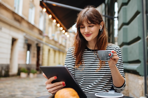 junge Frau mit braunen Haaren, die auf der Café-Terrasse sitzt und an einem digitalen Tablet arbeitet