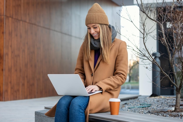 Junge Frau mit breitem Lächeln, die auf einer Bank sitzt, schaut auf Laptop mit Kaffee in ihren Händen auf Stadtstraße Manager im Nichtbüro ist mit zusätzlicher Arbeit am Laptop beschäftigt