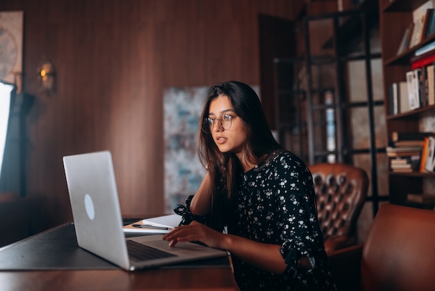 Junge Frau mit Brille arbeitet am Laptop, während sie am Tisch sitzt