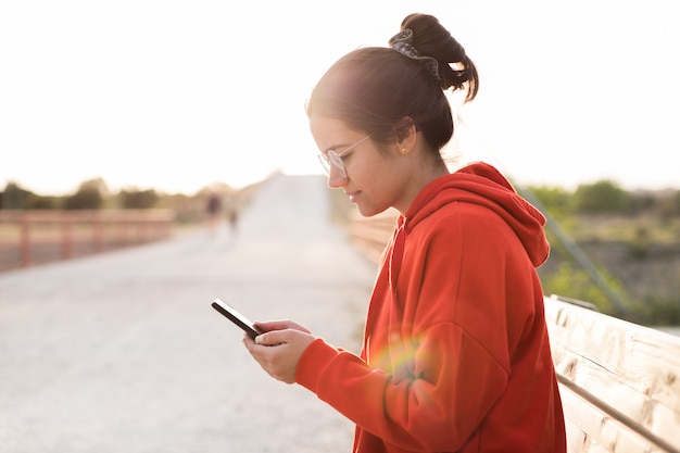 Junge Frau mit Brille auf ihren Zwanzigern, die auf ihrem Telefon plaudert, während sie in einer Bank eines Parks sitzt. Sie lächelt und trägt einen roten Pullover und einen Pferdeschwanz. Bei Sonnenuntergang ist sie von Tageslicht beleuchtet