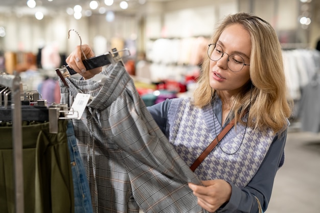 Junge Frau mit Brille, die beim Kauf von Kleidung im Laden eine neue Hose in den Händen betrachtet