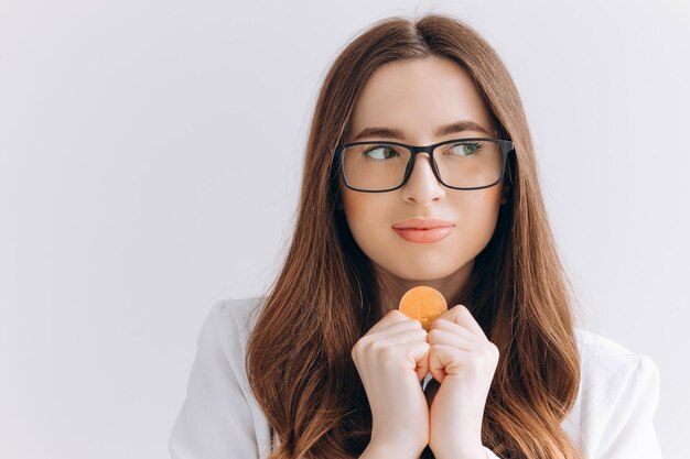 Junge Frau mit Brille, die einen Bitcoin isoliert auf grauem Hintergrund hält und Geldgeste macht