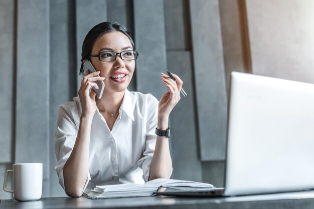 Junge Frau mit Brille, die im Büro am Laptop arbeitet, während sie telefoniert