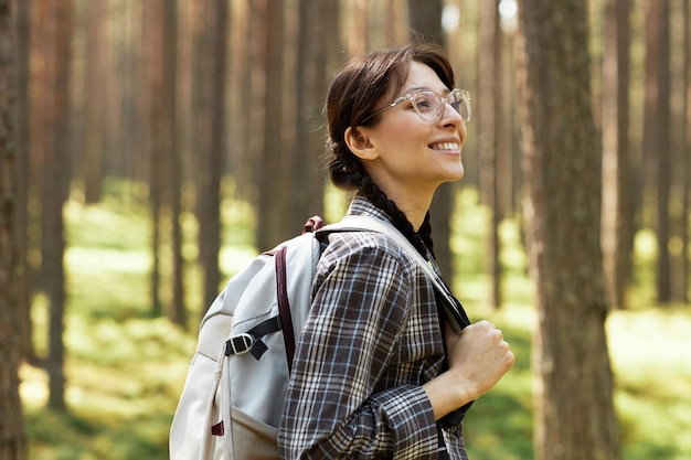 Junge Frau mit Brille und Rucksack hinter dem Rücken genießt die Natur beim Wandern im Wald