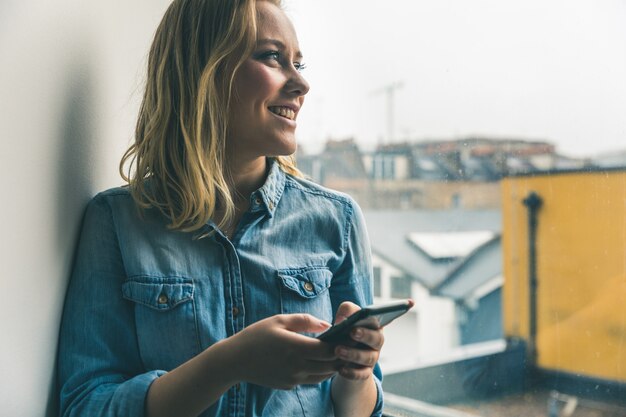 Junge Frau mit dem Telefon, das aus Fenster heraus schaut