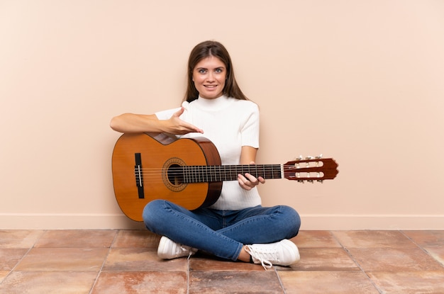 Junge Frau mit der Gitarre, die auf den ausdehnenden Händen des Bodens zur Seite für die Einladung sitzt zu kommen