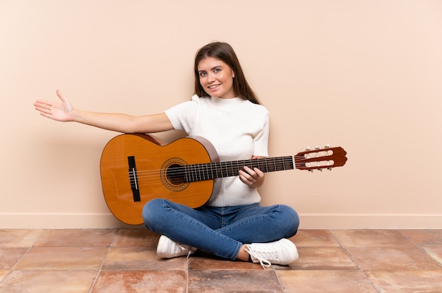 Junge Frau mit der Gitarre, die auf den ausdehnenden Händen des Bodens zur Seite für die Einladung sitzt zu kommen