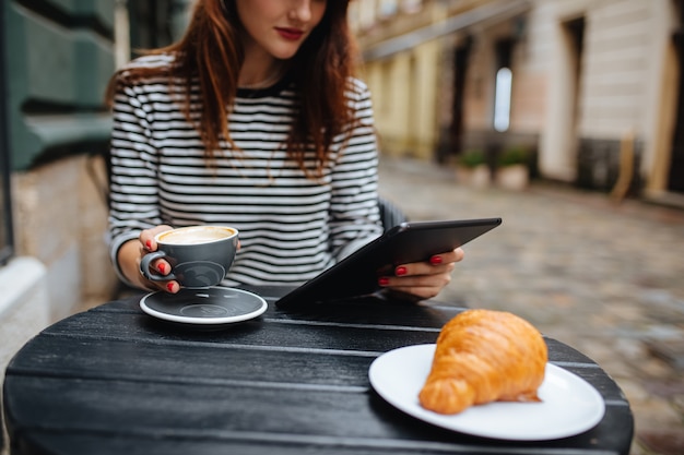 junge Frau mit digitalem Tisch mit leckerem Kaffee und Croissant auf der Café-Terrasse