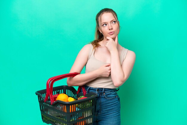 Junge Frau mit einem Einkaufskorb voller Lebensmittel isoliert auf grünem Hintergrund mit Zweifeln