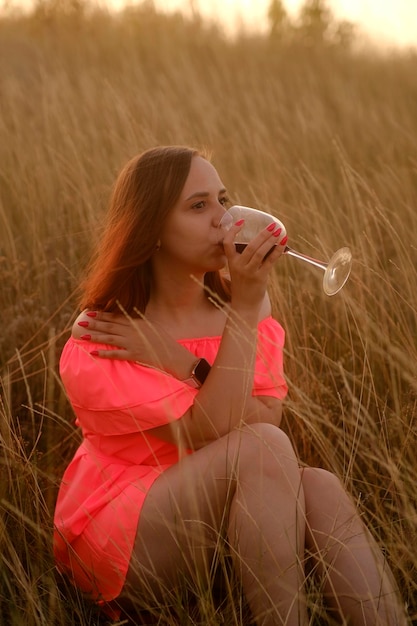 Junge Frau mit einem Glas Wein auf dem Feld Seitenansicht einer jungen Frau in orangefarbenem Kleid, die Rotwein trinkt und wegschaut, während sie auf dem Gras auf dem Land sitzt