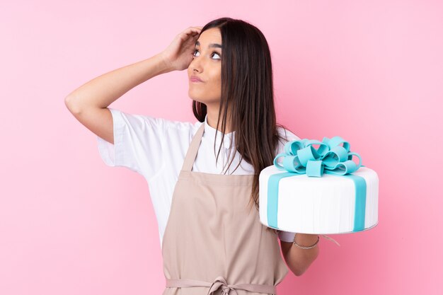 Junge Frau mit einem großen Kuchen über isolierter Wand, die Zweifel und mit verwirrendem Gesichtsausdruck hat