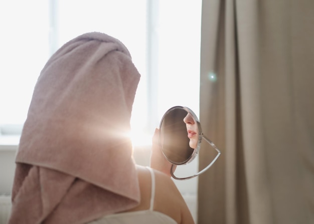 Junge Frau mit einem Handtuch auf dem Kopf trägt Creme auf die Gesichtshaut auf und schminkt sich zu Hause