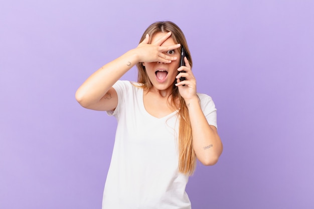 Junge Frau mit einem Handy, das schockiert, verängstigt oder verängstigt aussieht und das Gesicht mit der Hand bedeckt