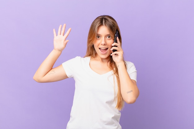 junge Frau mit einem Handy, die glücklich lächelt, die Hand winkt, Sie begrüßt und begrüßt