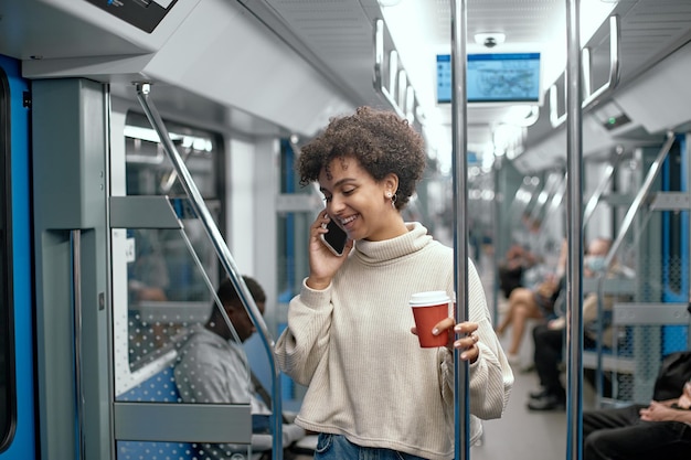 Junge Frau mit einem Kaffee zum Mitnehmen, der auf einem Smartphone in einem U-Bahn-Wagen spricht