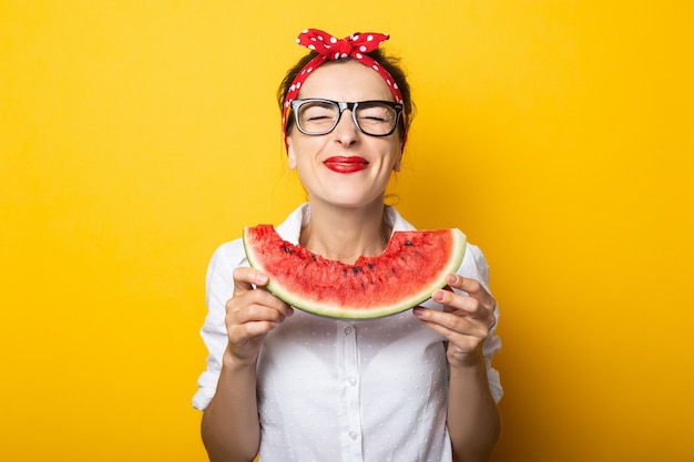Junge Frau mit einem Lächeln in einem roten Stirnband und Gläsern isst eine Wassermelone an einer gelben Wand.