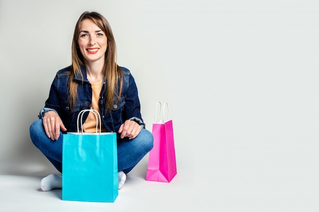 Junge Frau mit einem Lächeln in einer Jeansjacke mit Einkaufstüten beim Sitzen auf dem Boden auf einem hellen Raum. Banner. Konzept einkaufen, verkaufen.