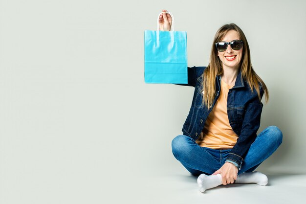 Junge Frau mit einem Lächeln in einer Jeansjacke und einer Sonnenbrille hält Einkaufstaschen, während sie auf dem Boden auf einem hellen Raum sitzt.