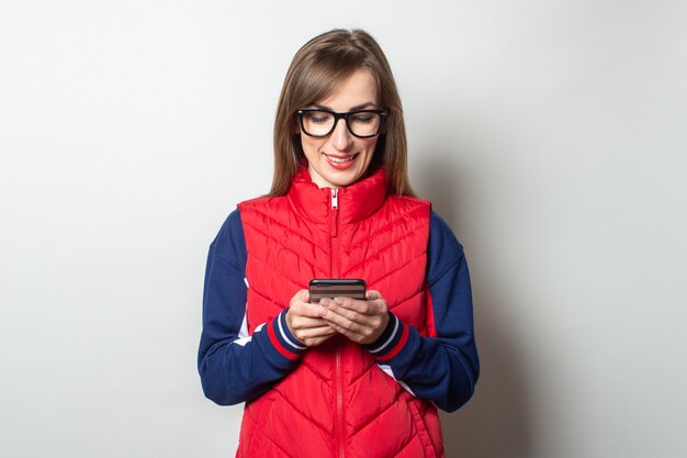 Junge Frau mit einem Lächeln in einer roten Weste schaut in ihr Telefon gegen eine helle Wand