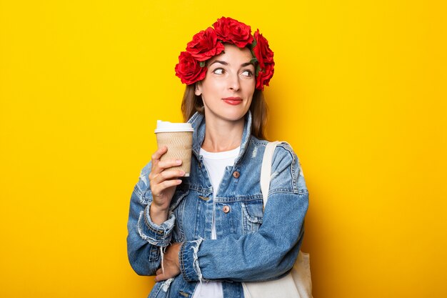 Junge Frau mit einem Lächeln schaut zur Seite in einer Jeansjacke und ein Kranz aus roten Blumen auf ihrem Kopf hält eine Pappbecher mit Kaffee an einer gelben Wand.