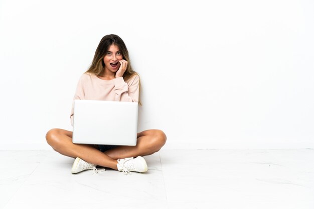 Junge Frau mit einem Laptop, der auf dem Boden lokalisiert auf Weiß mit Überraschung und schockiertem Gesichtsausdruck sitzt
